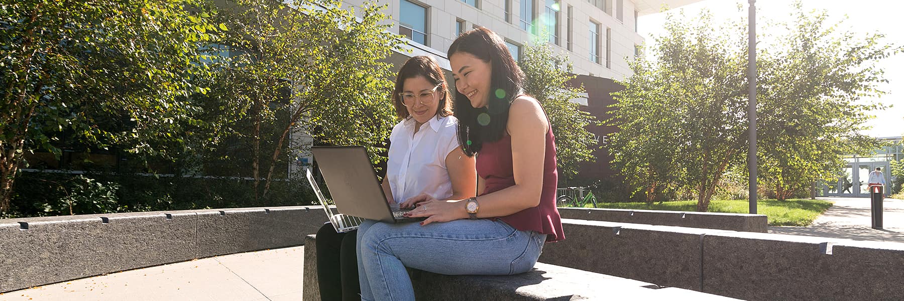 2 linguistics grad students look at laptop outside University Hall on a bench.